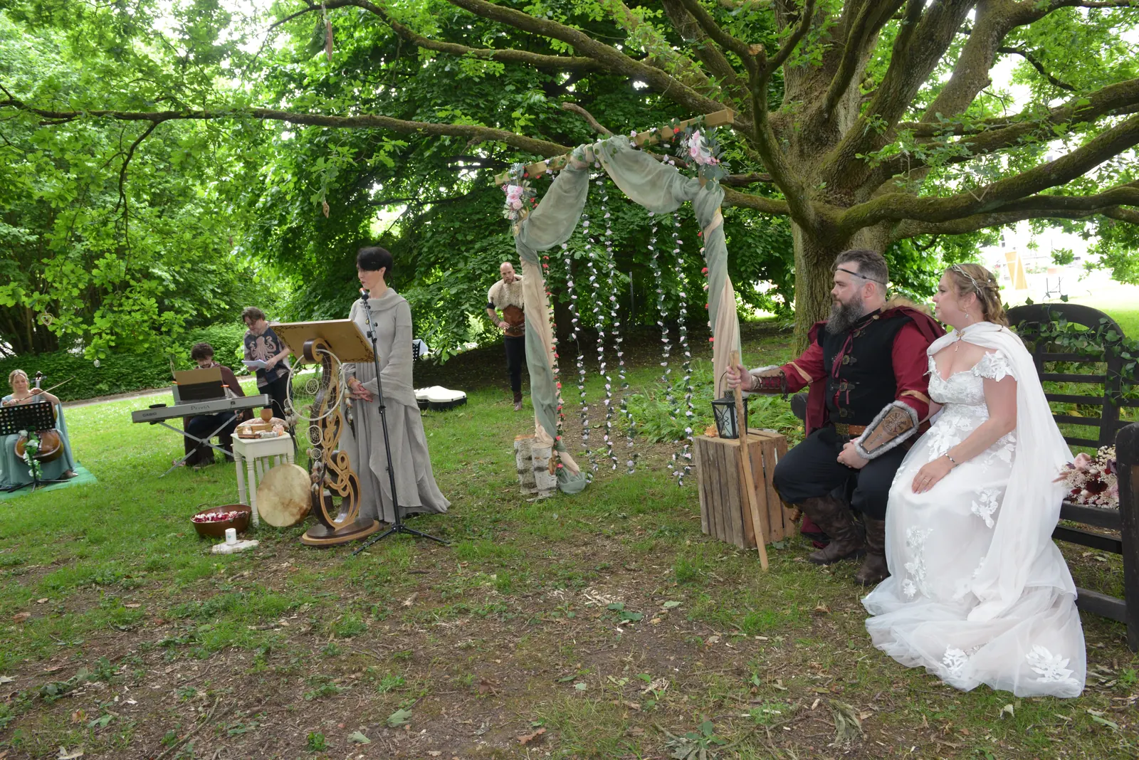 Espace de cérémonie viking sous un arbre centenaire : pupitre sculpté, mariés sur un banc et officiante